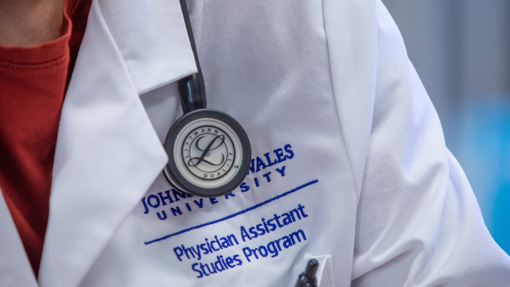 Closeup of a stethoscope and a white coat embroidered with the words, “Johnson & Wales University Physician Assistant Studies Program.”
